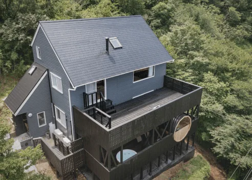 森に囲まれた青い外壁の建物とウッドデッキを見下ろす空撮