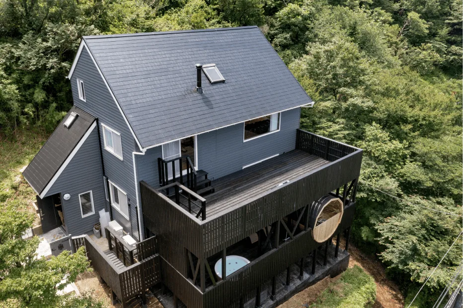 緑に囲まれた青い屋根の建物と黒いウッドデッキ（バレルサウナ・ジャグジー）の空撮