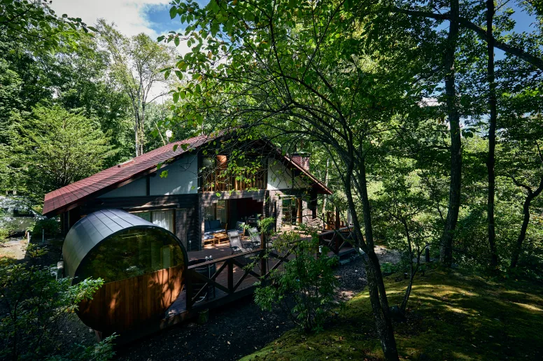 森の木々に囲まれた建物とウッドデッキのバレルサウナ